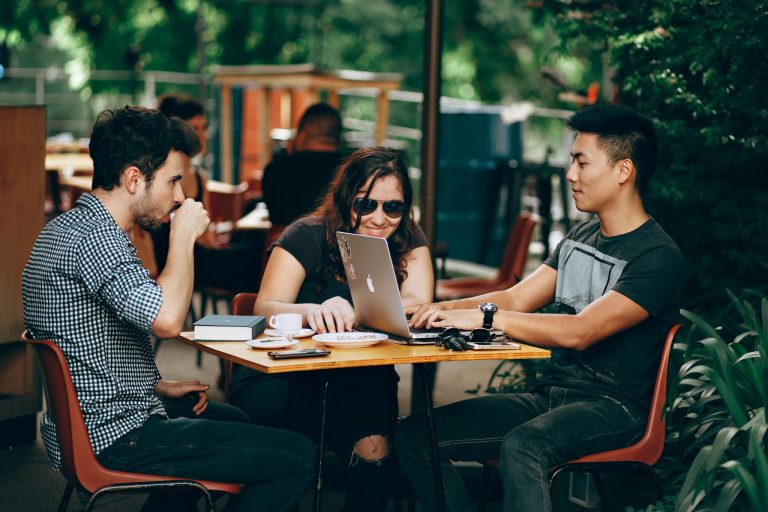 A group of young adults working on a laptop at an outdoor coffee shop, enjoying teamwork and collaboration.